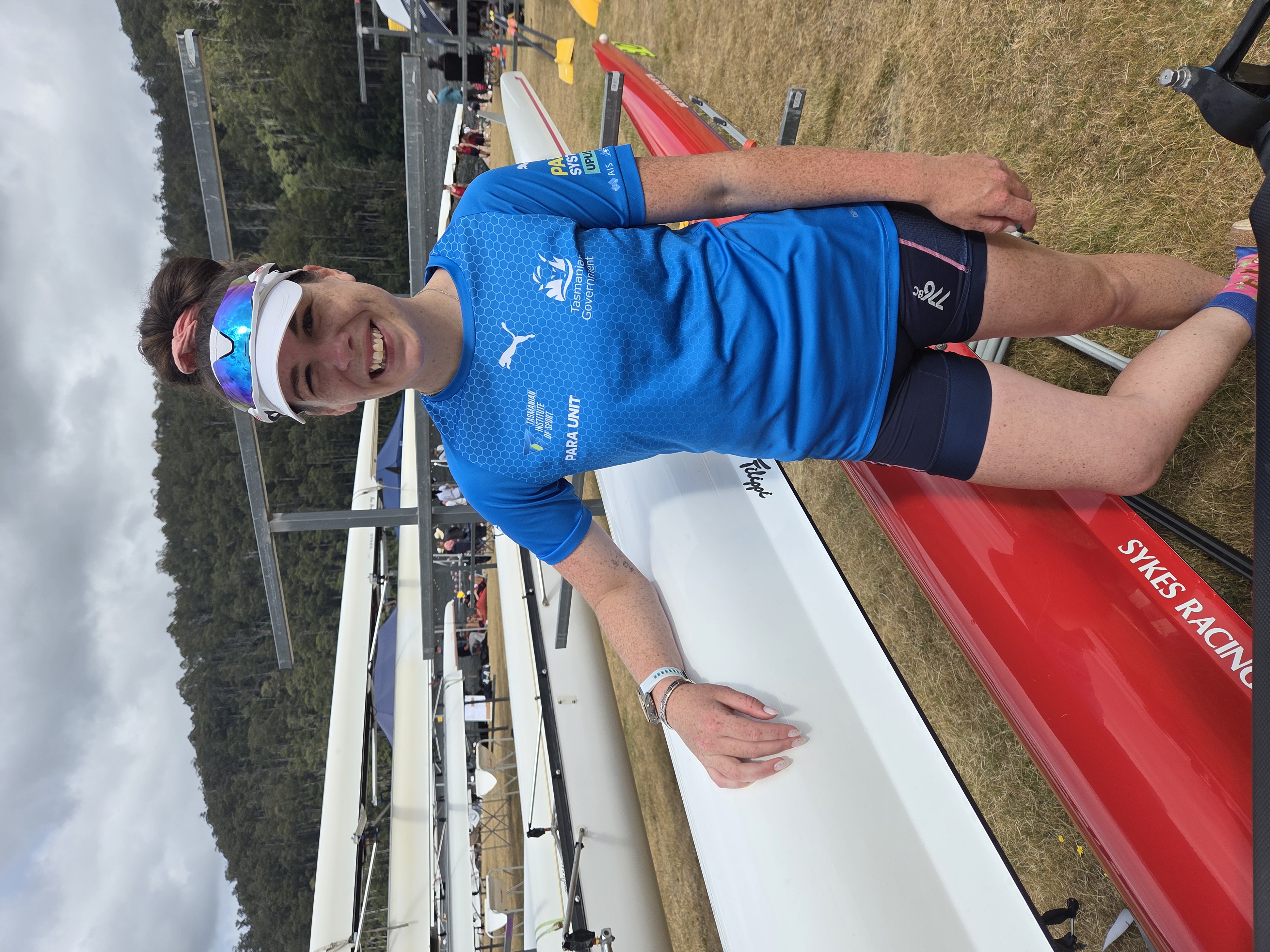 Ella Marshall is photographed in her TIS Para Unit training uniform leaning against rowing shells after a race at Lake Barrington